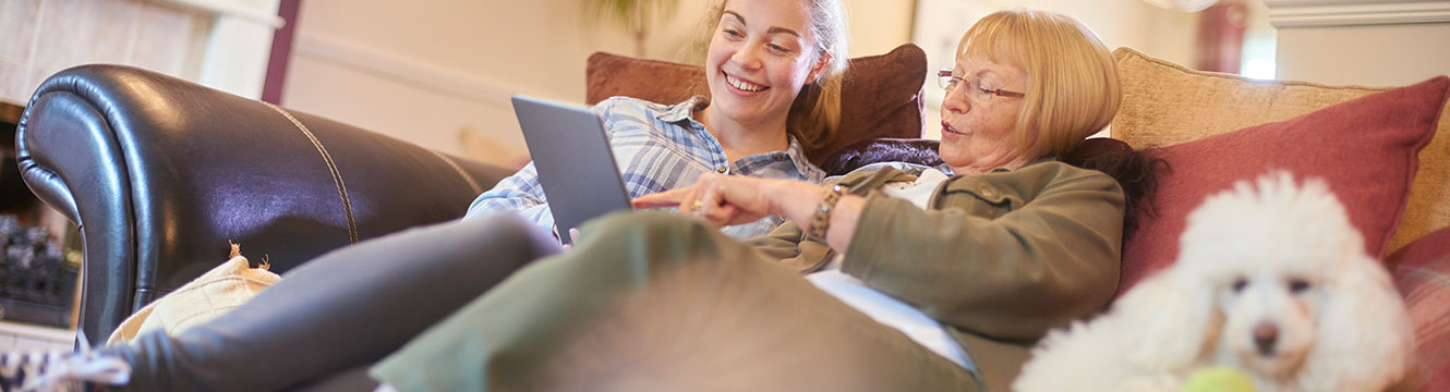 Girl and her grandmother looking at tablet while sitting on the couch with small poodle dog