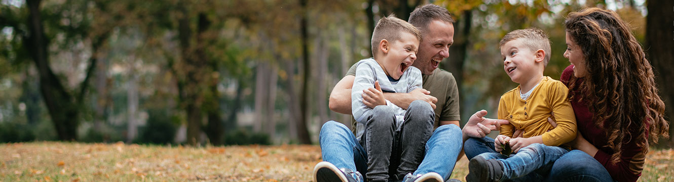 Mom holding one son smiling and laughing at father holding the other son sitting outside on the ground in the leaves