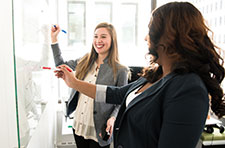 Two women writing on a wall-sized white board